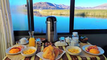 A breakfast table with various food items and a coffee maker, set against a scenic view of a lake and mountains.