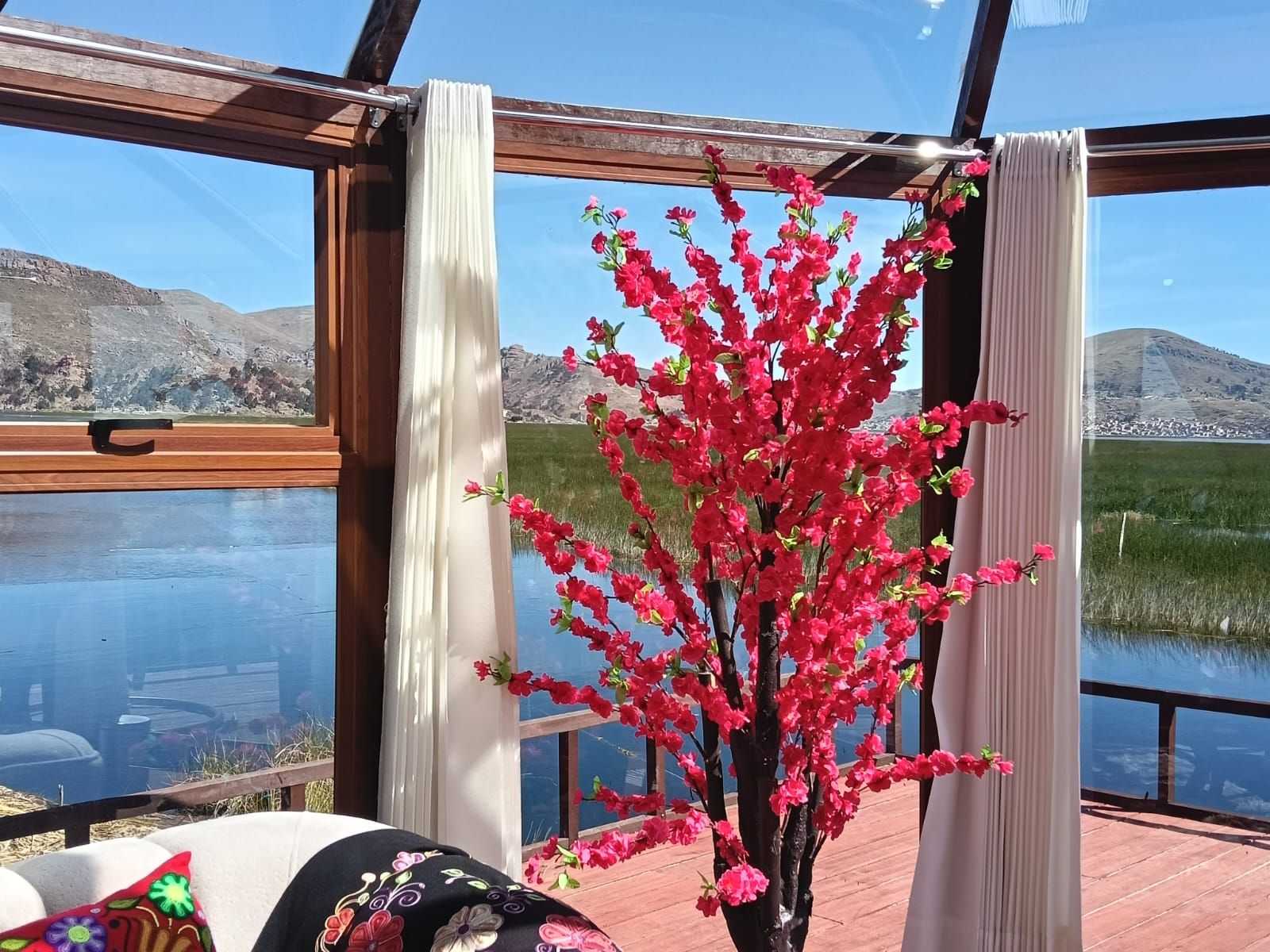 Una habitación con un gran ventanal, una mesa de madera y una planta en maceta con flores rosadas.