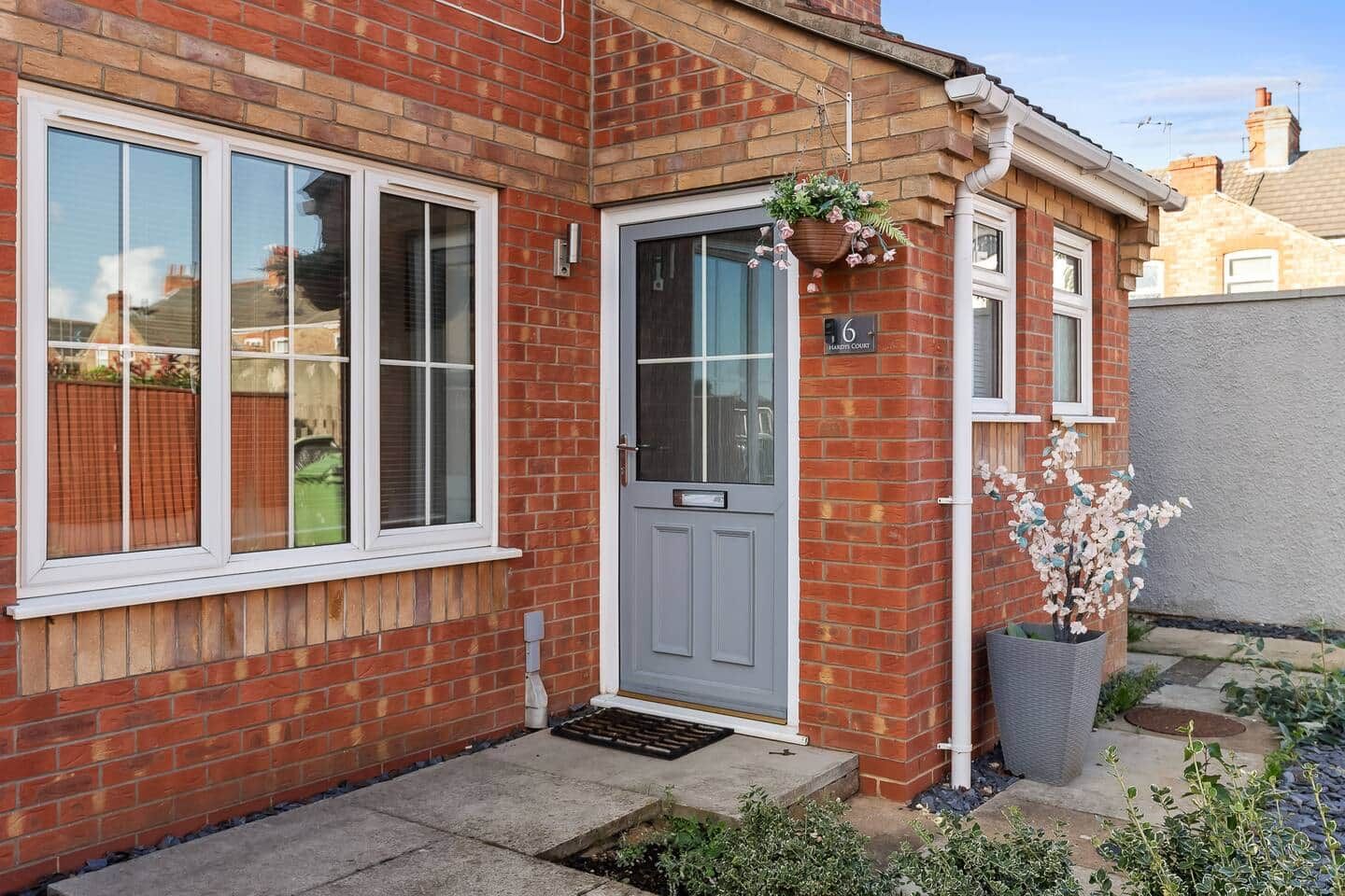 Une maison en briques avec une porte grise, des fenêtres à cadre blanc et un petit jardin agrémenté d’une plante en pot.