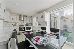 Een moderne keuken met witte kastjes, een eettafel met een bloemrijk tafelkleed en een groot raam met jaloezieën.