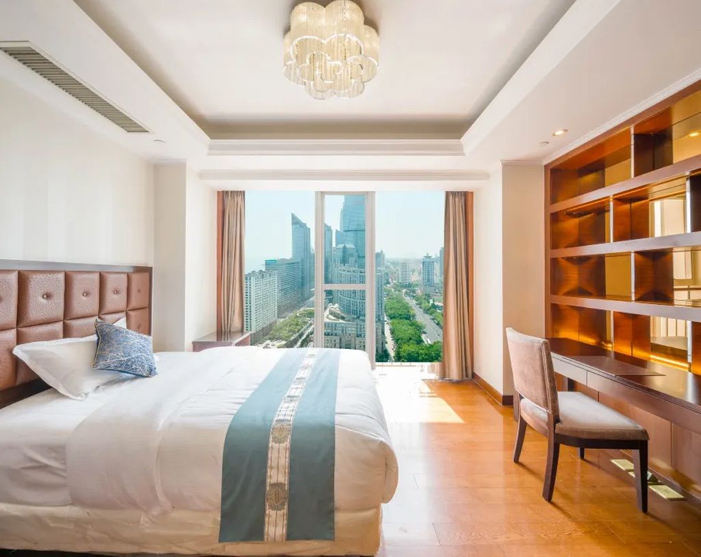 A hotel room with a large bed, a view of the cityscape, a bookshelf, and a desk.