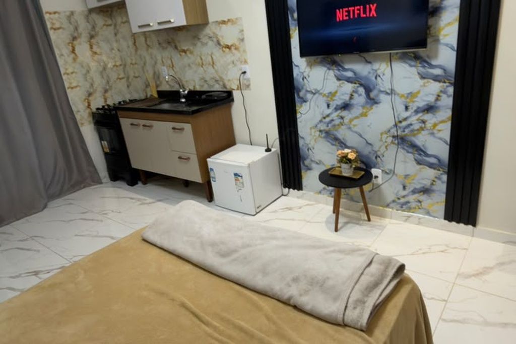 A hotel room with a bed, a small table with a vase, a television displaying 'NETFLIX', and a kitchen area.