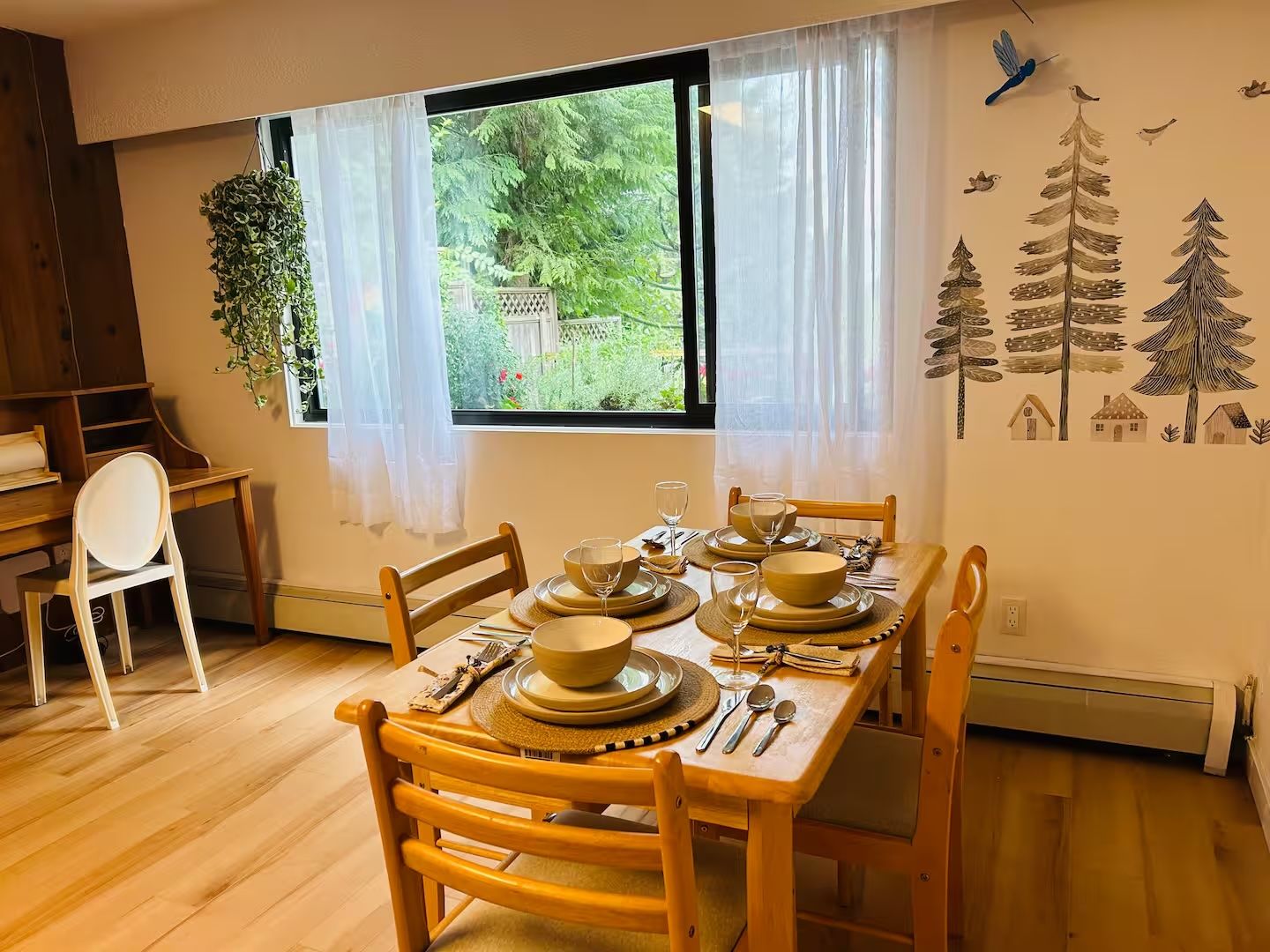 Un coin repas avec une table dressée pour quatre personnes, une fenêtre donnant sur un paysage verdoyant et un mur décoré d’autocollants représentant des arbres et des maisons.