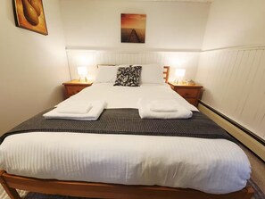 A neatly made bed with white linens and a striped blanket, two bedside tables with lamps, and a sunset painting on the wall. - Private suite near trails with mountain view (North Vancouver)