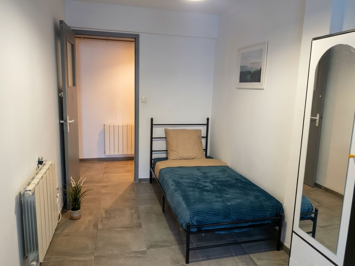 A small, single-bed room with a radiator, a potted plant, and a framed picture on the wall.