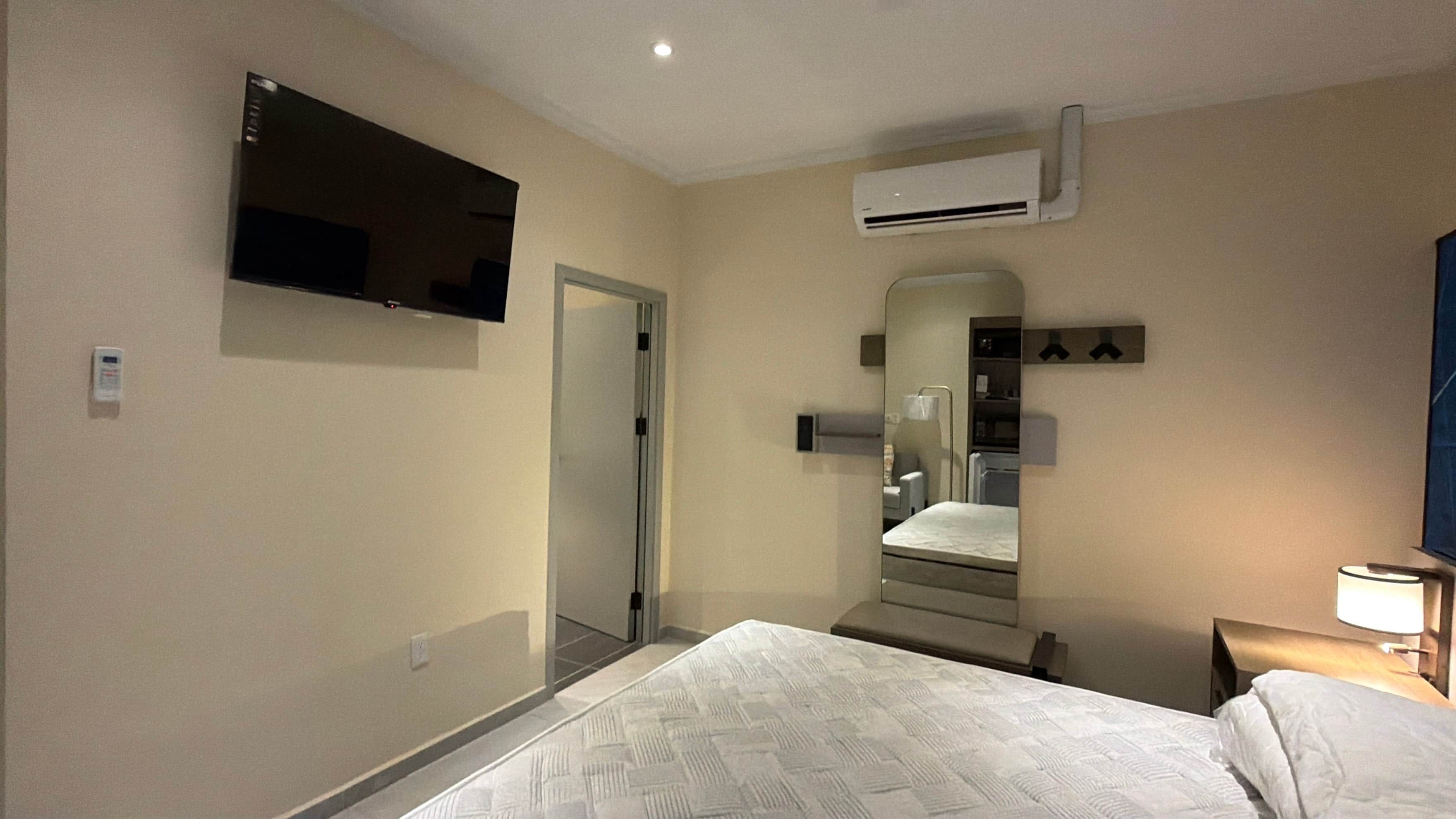 Habitación de hotel con cama, televisor de pantalla plana montado en la pared, aire acondicionado, mesita de noche con lámpara y espejo.