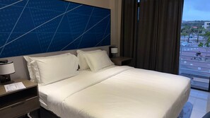 Habitación de hotel con una cama grande, una mesita de noche, una lámpara y vistas a la ciudad desde la ventana.