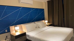 Habitación de hotel con una cama grande, dos mesitas de noche con lámparas, un cabecero con diseño geométrico y una ventana con cortinas.
