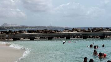 Escena costera con un muelle que se adentra en el agua, rodeado de rocas y una vista distante de una colina y estructuras industriales.