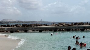 Escena costera con un muelle que se adentra en el agua, rodeado de rocas y una vista distante de una colina y estructuras industriales.