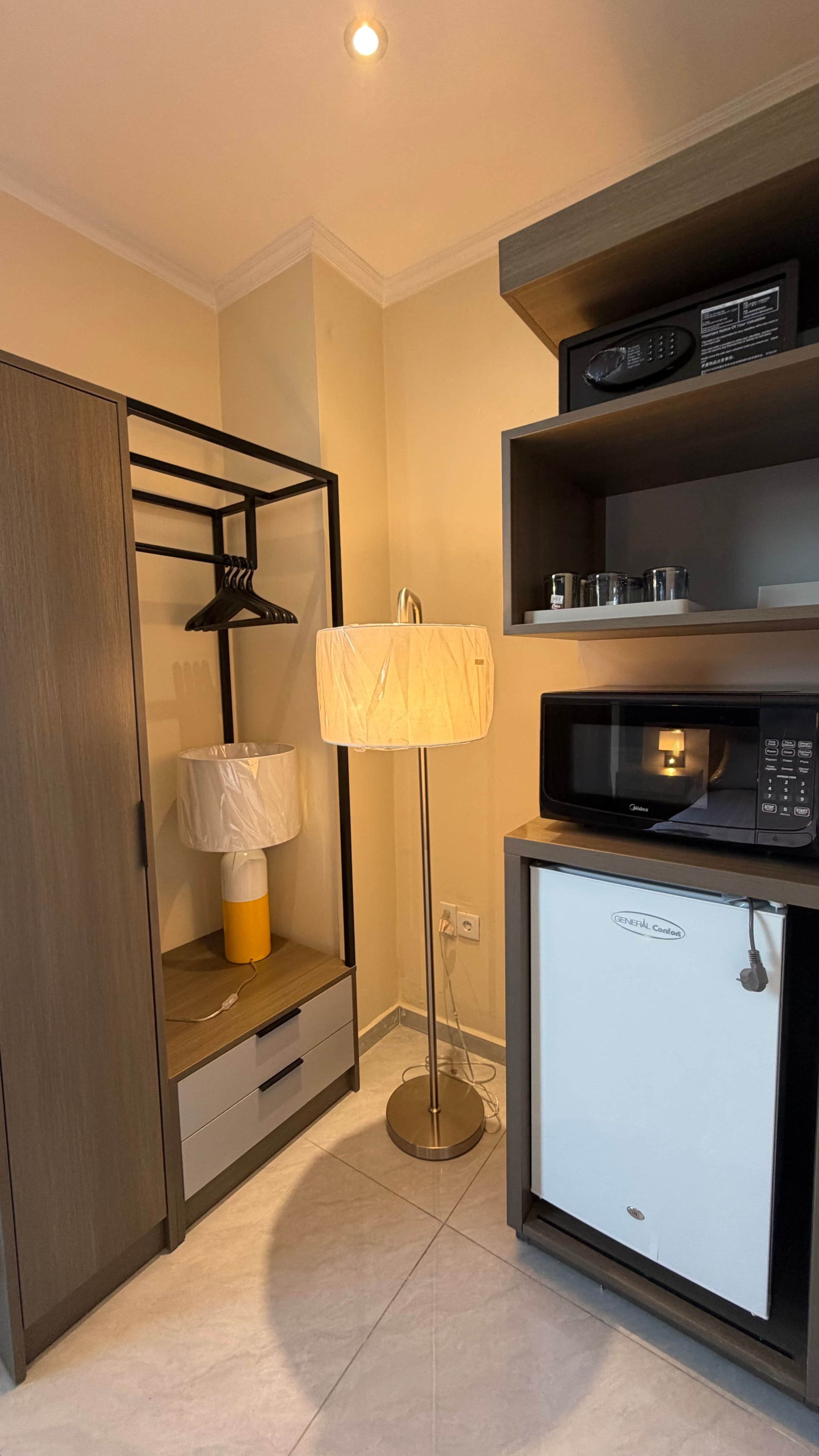 Habitación de hotel compacta con armario, lámpara de pie, microondas y refrigerador pequeño.