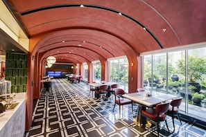 Un restaurante con una decoración moderna, que presenta un suelo estampado, techo curvo rojo y grandes ventanales con vistas a un jardín.