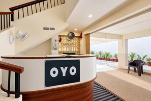 Uma área de recepção com balcão de madeira identificado com a marca 'OYO', um relógio e vista para o exterior através de grandes janelas.