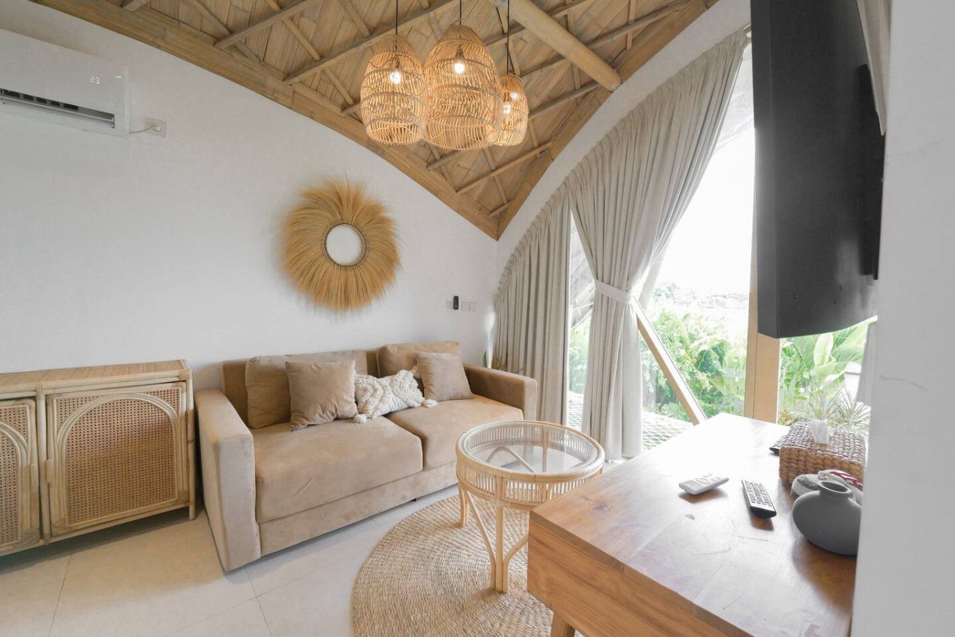 A room with a wooden ceiling, a rattan sofa, a round table, and a view of greenery outside.