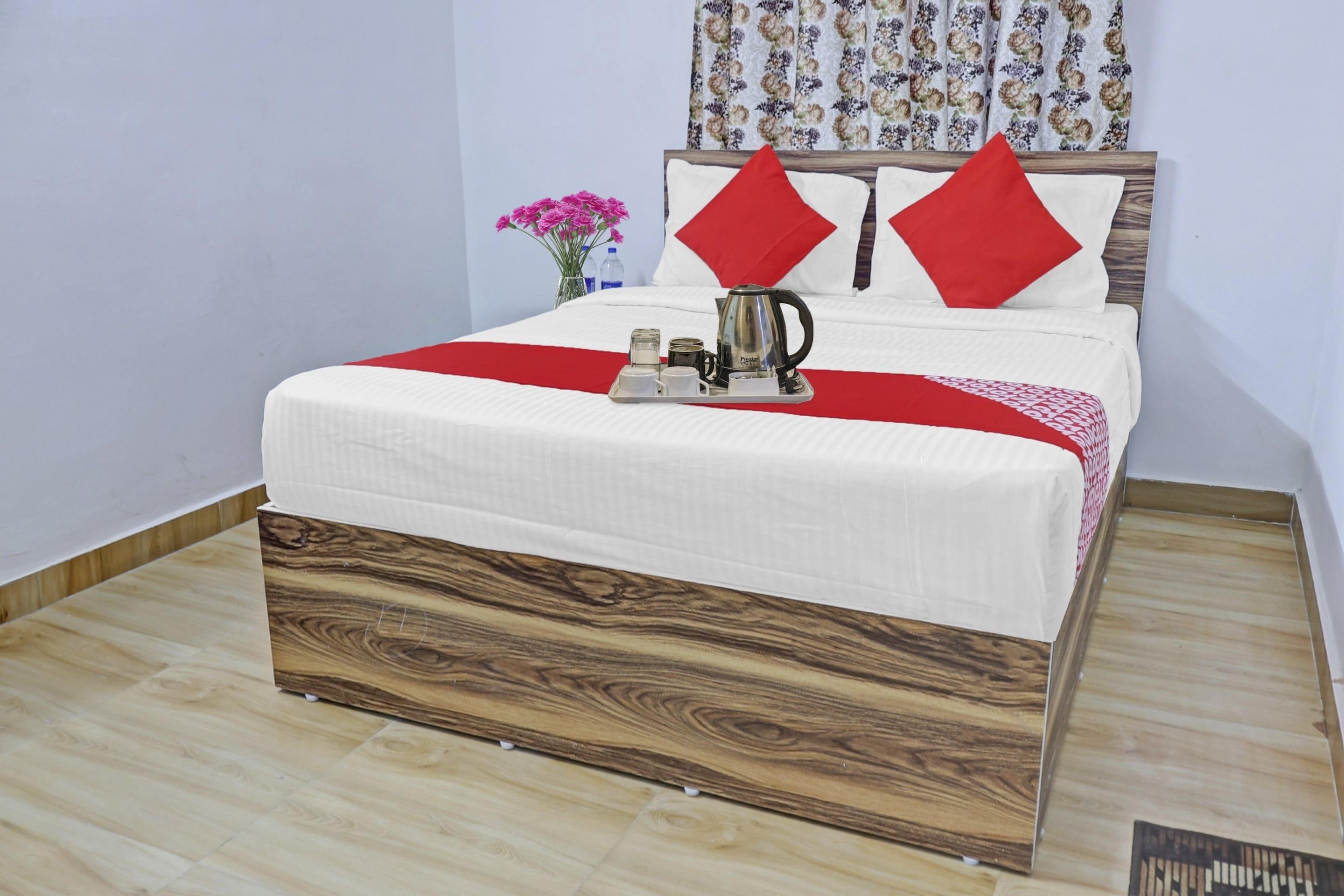 A neatly made bed with a wooden headboard, a red throw blanket, and a tray with a kettle and cups.