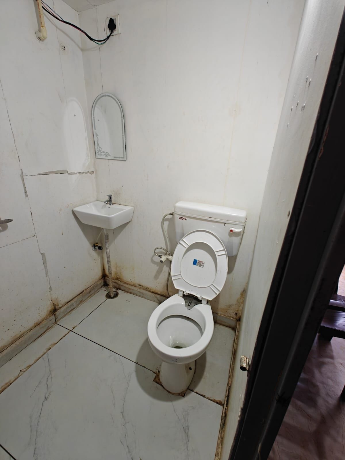 A small, compact bathroom with a white toilet, a sink, and a mirror. The walls and floor are tiled, and the space is enclosed by a door.