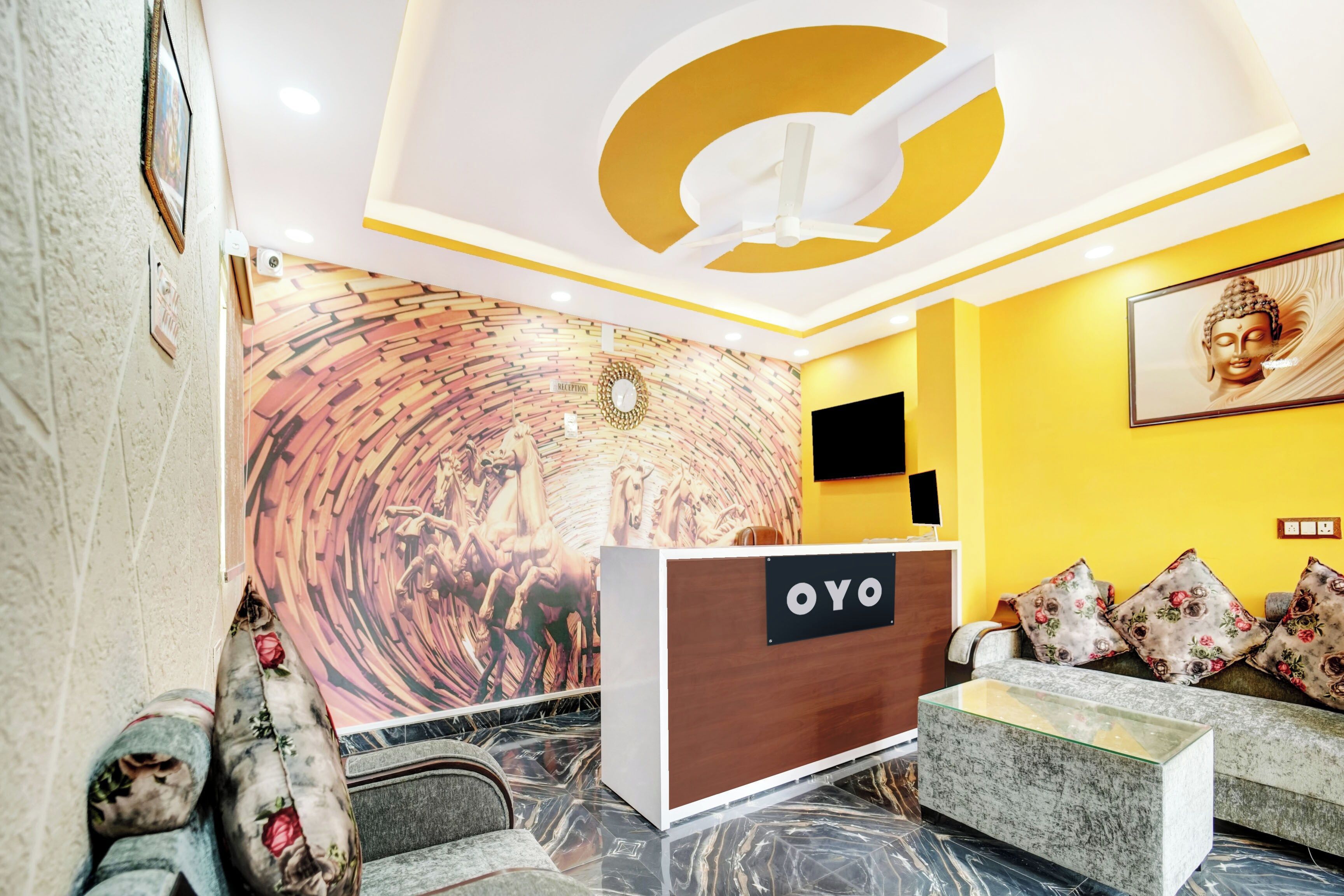 A reception area with a reception desk labeled 'OYO,' a wall mural, a Buddha statue, and a sofa with floral cushions.