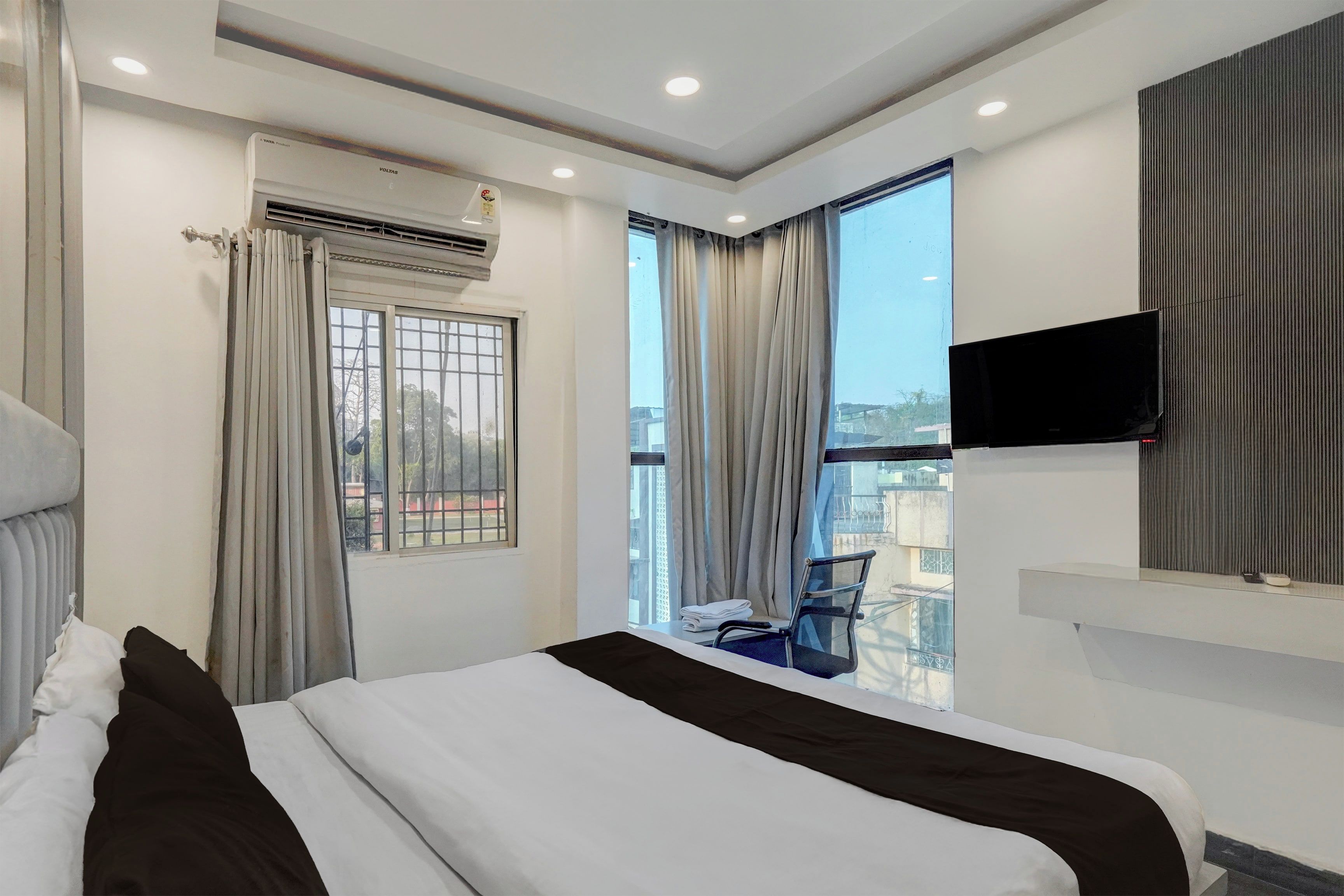 A hotel room with a bed, a flat-screen TV mounted on the wall, a window with curtains, and a view of the outside.