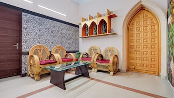 Una habitación con una mesa de vidrio, cuatro sillas de mimbre con cojines rojos, una puerta de madera con intrincados grabados y un papel tapiz estampado.