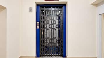 Puerta de ascensor azul con una reja metálica, un pequeño cartel encima y piso de baldosas.