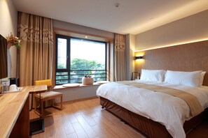 Quarto de hotel com uma cama grande, uma mesa com cadeira, uma janela com cortinas e vista para o verde lá fora.