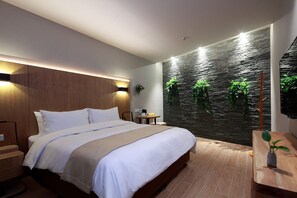 Quarto de hotel moderno com cama grande, cabeceira de madeira, TV na parede e uma pequena mesa com uma planta.