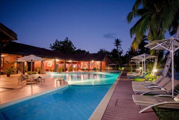 A swimming pool with lounge chairs and umbrellas, surrounded by palm trees and a building with a thatched roof. - Mantis Soanambo Hotel and Spa Sainte Marie (Nosy Boraha)