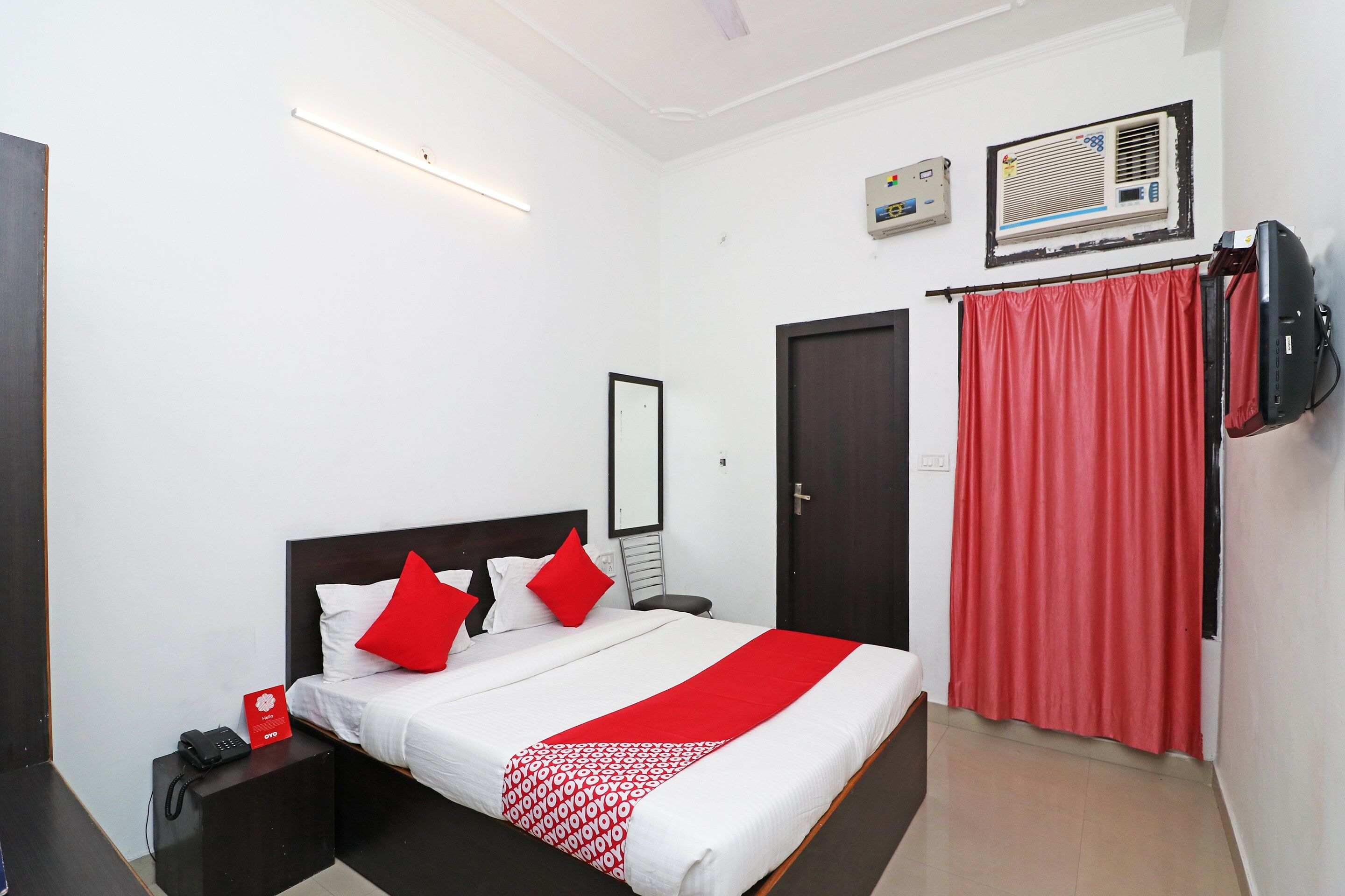 Habitación de hotel con una cama, un teléfono en la mesita de noche, una colcha de cuadros blancos y rojos, una cortina roja y un televisor montado en la pared.