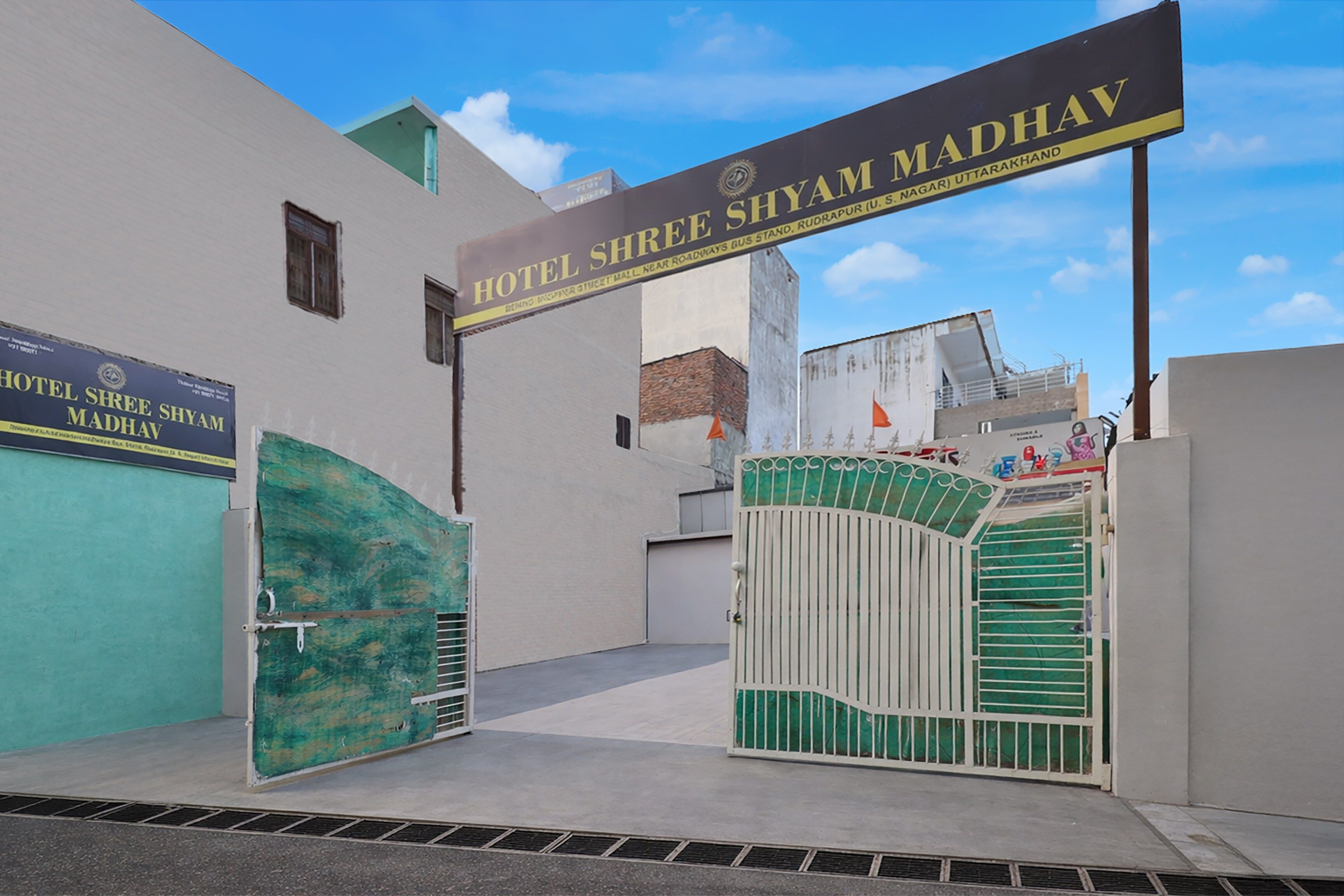 Entrada de un hotel con un letrero que dice 'Hotel Shree Shyam Madhav' y un camino de acceso con portones.