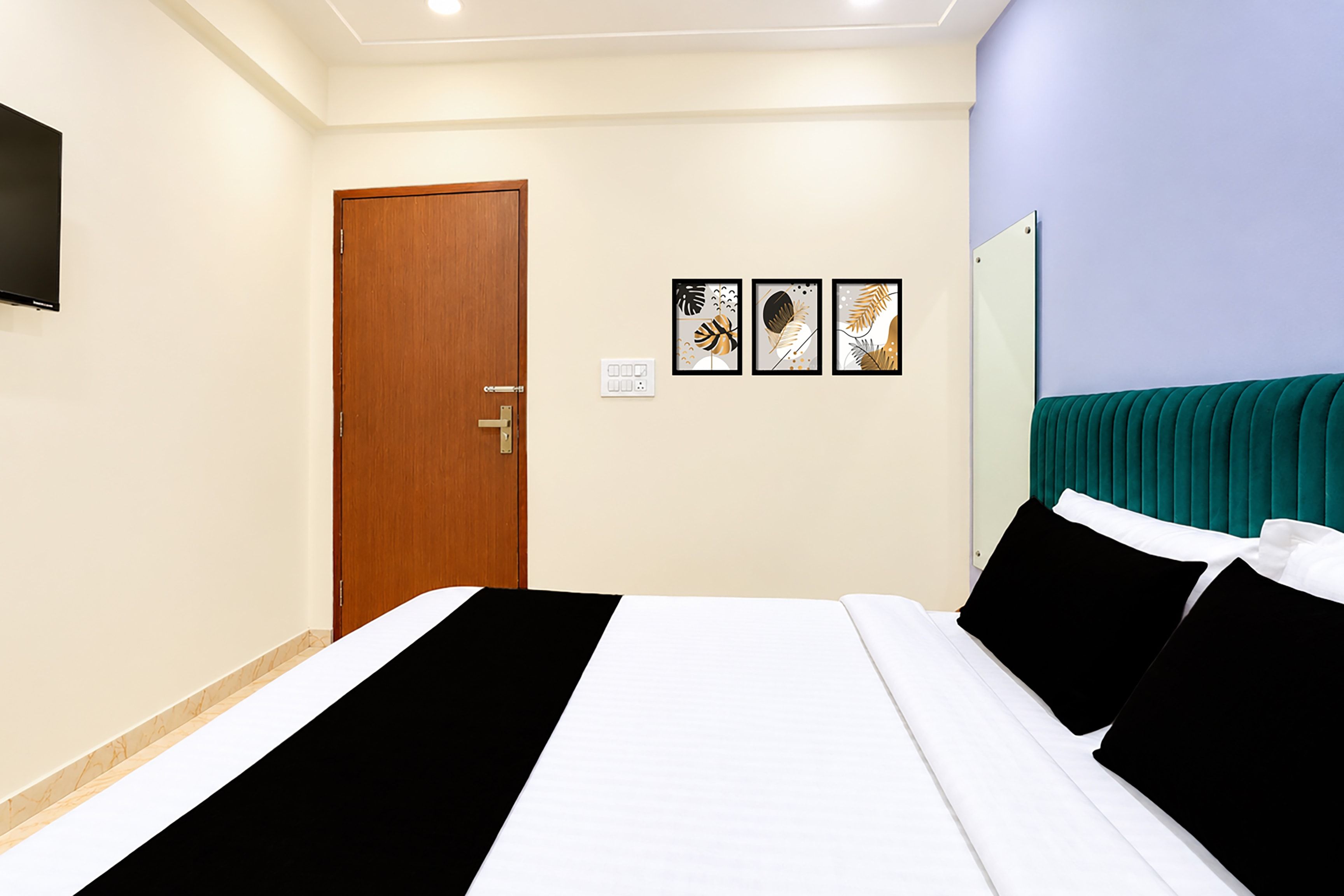 A hotel room with a bed, a door, a wall-mounted TV, and three framed artworks.