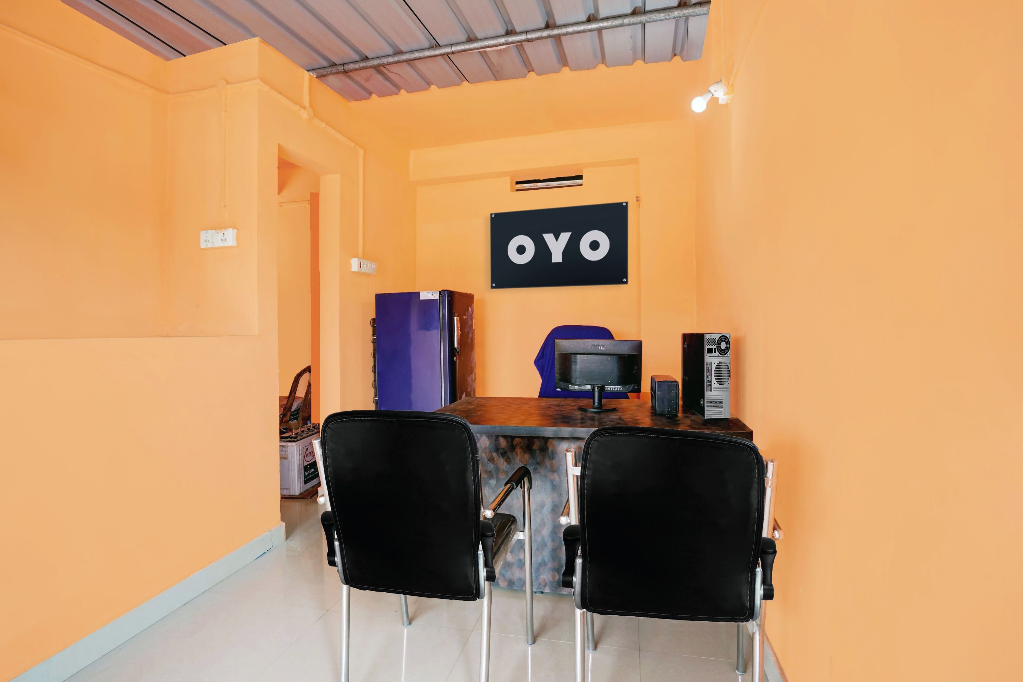 A small, orange-walled room with a desk, two chairs, a refrigerator, a computer, and a sign that reads 'OYO'.