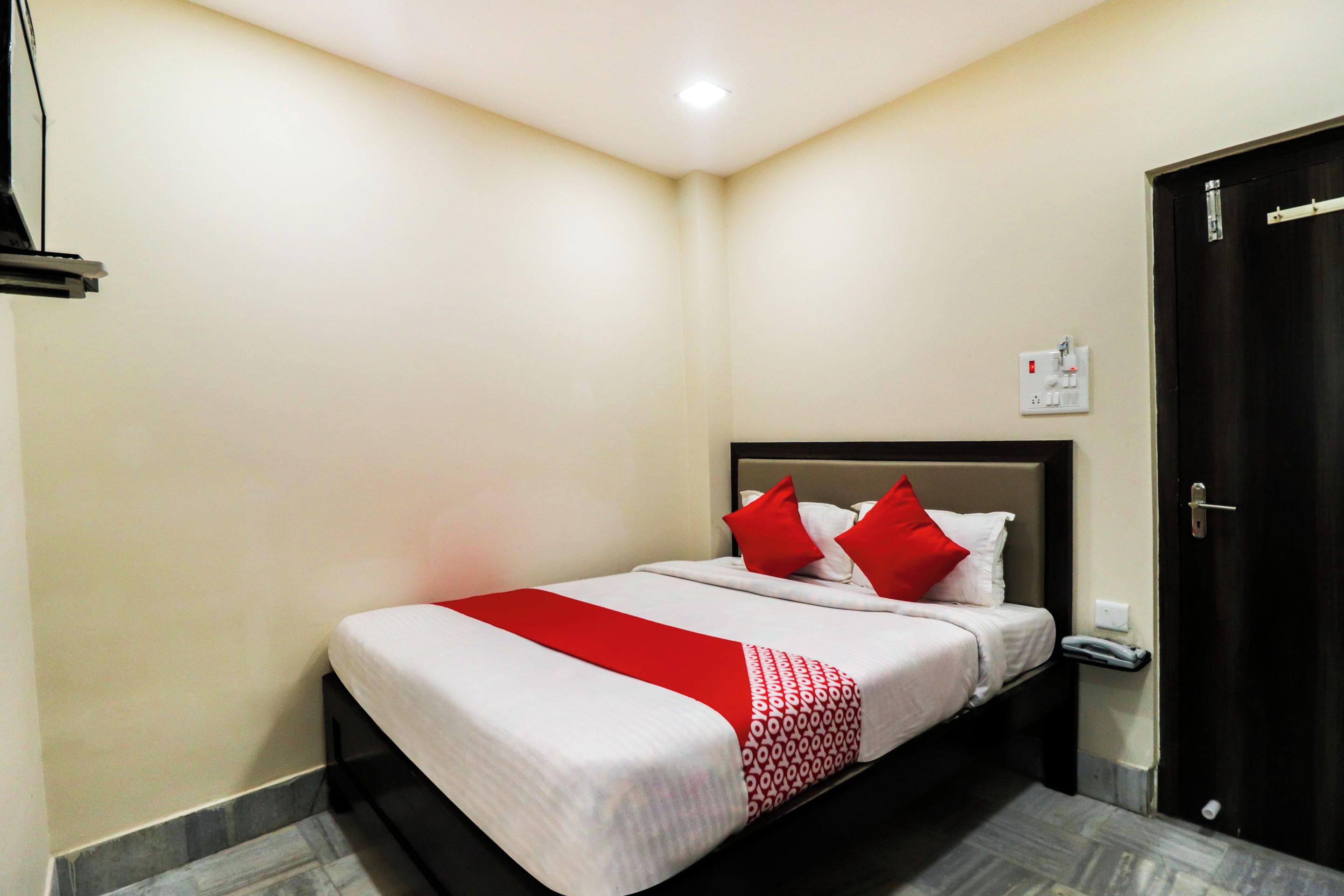 A hotel room with a bed, a red and white bedspread, a black headboard, a white wall, a door, and a light switch.
