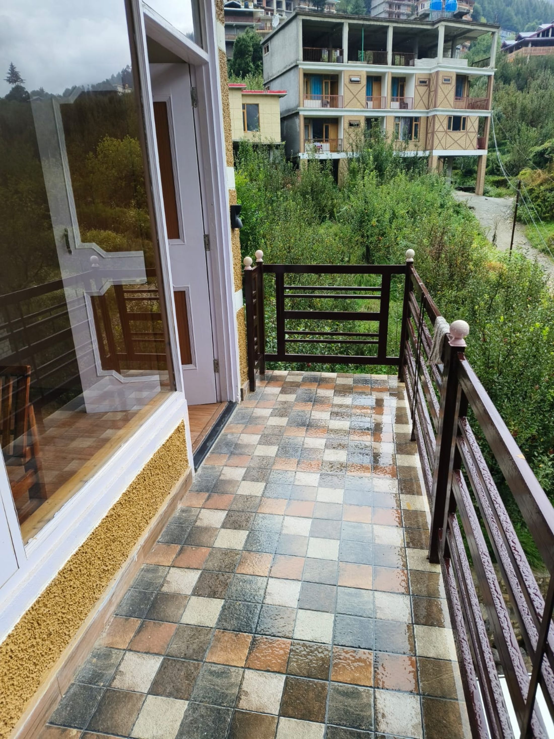 A balcony with a tiled floor leading to a wooden gate, surrounded by greenery and residential buildings.