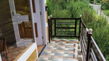 A balcony with a tiled floor leading to a wooden gate, surrounded by greenery and residential buildings.