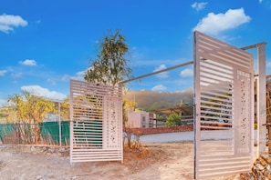 Uma entrada gradeada com portão metálico e painel decorativo, conduzindo a um edifício com telhado de azulejos e vista para uma paisagem acidentada.