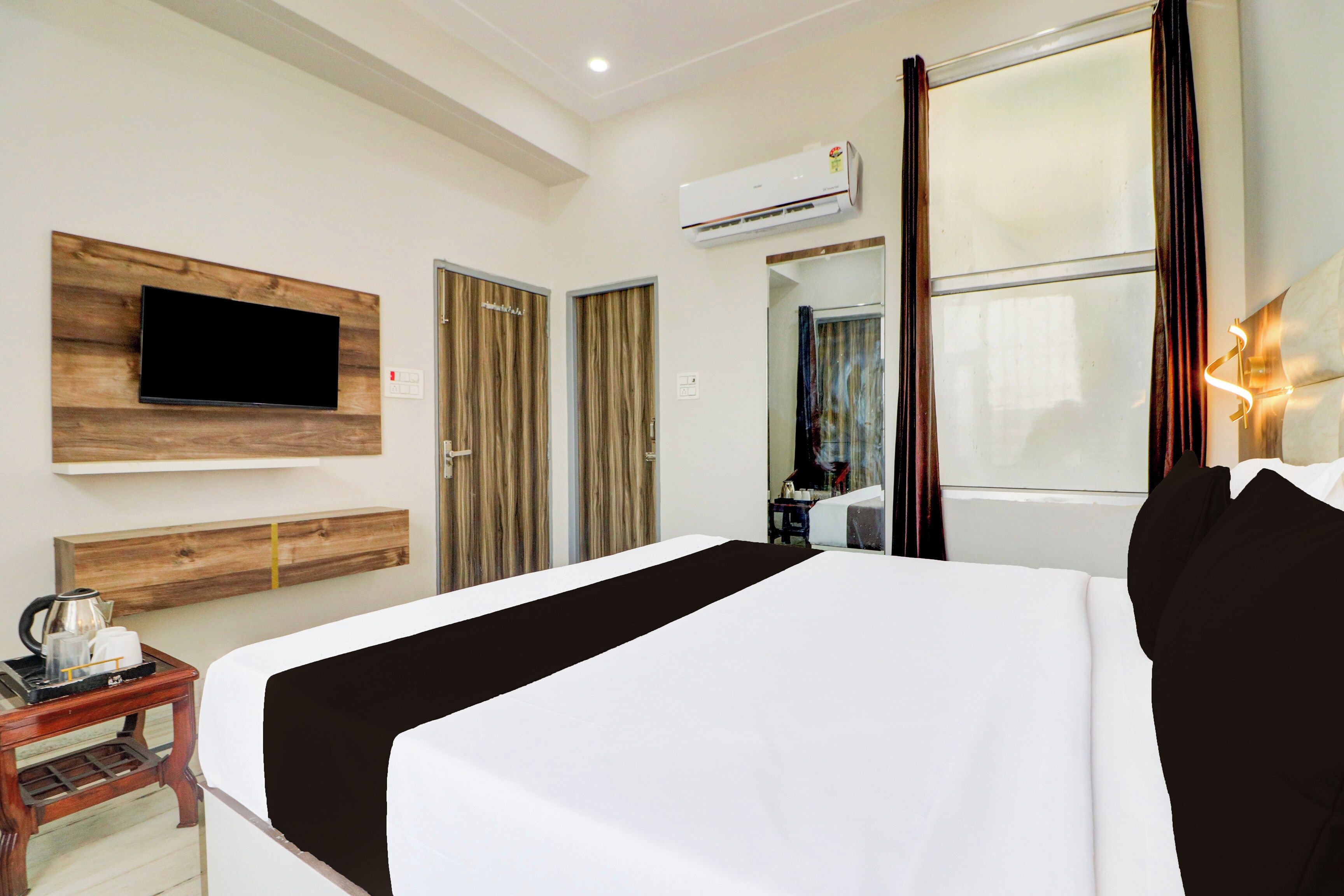 A hotel room with a bed, a flat-screen TV mounted on a wooden panel, a coffee maker on a side table, and an air conditioning unit on the wall.