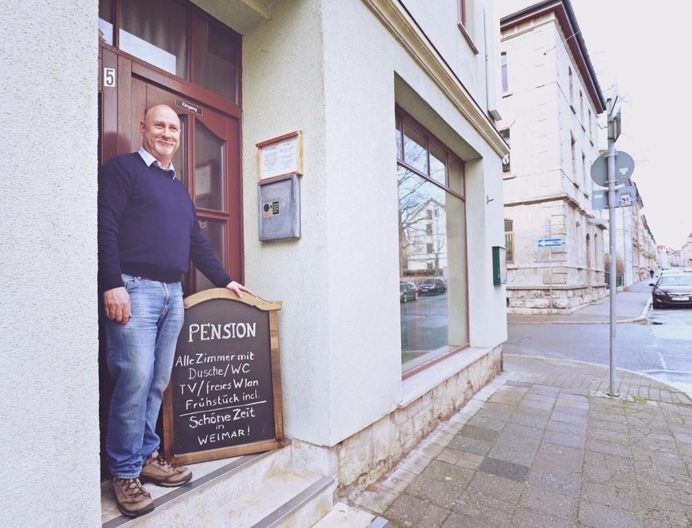 Un homme se tient devant un immeuble, et une enseigne annonce une pension proposant des chambres équipées d’une télévision, d  ’une connexion Wi-Fi gratuite et d’un réfrigérateur.