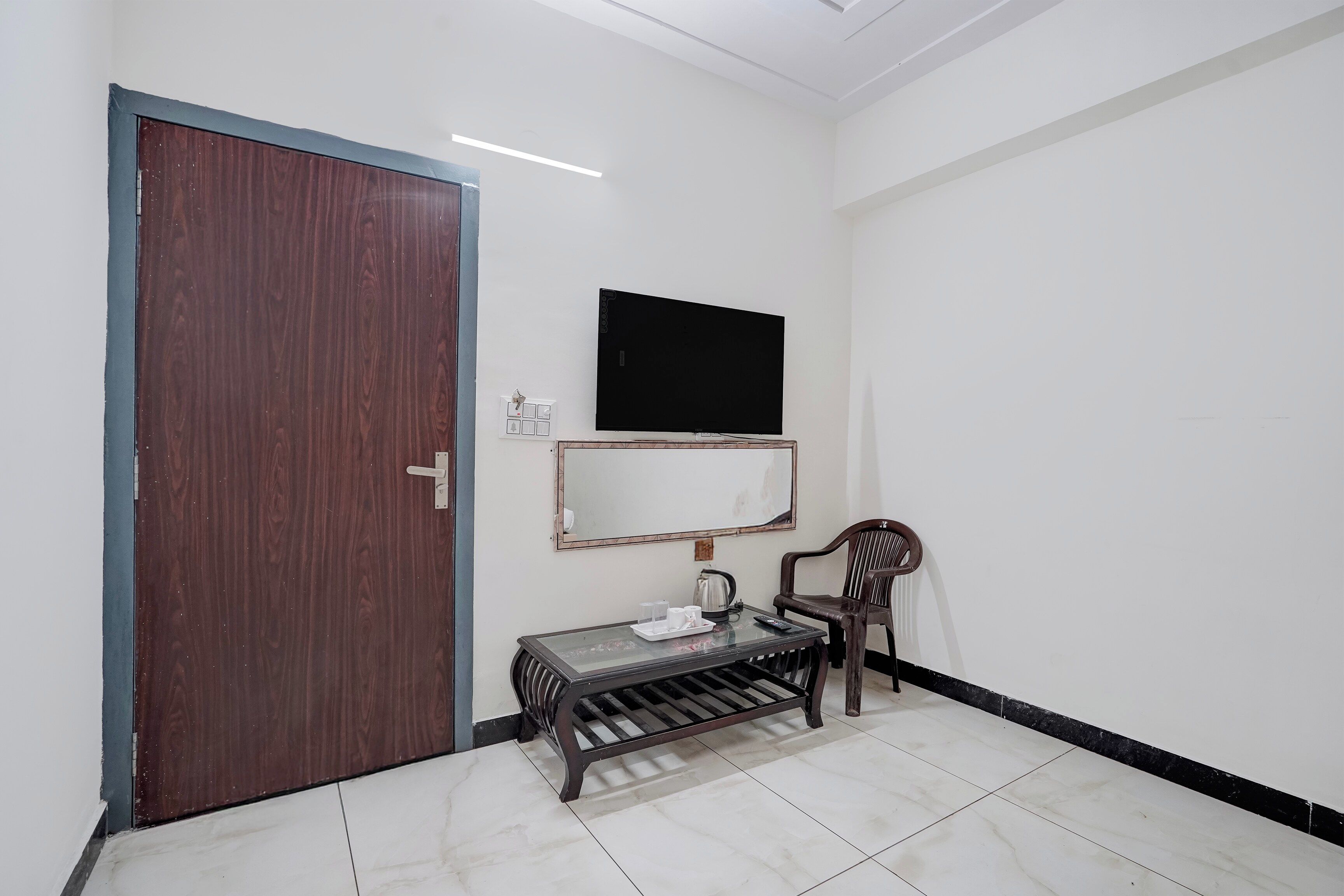 A room with a wooden door, a television mounted on the wall, a small table with a chair, and a tiled floor.