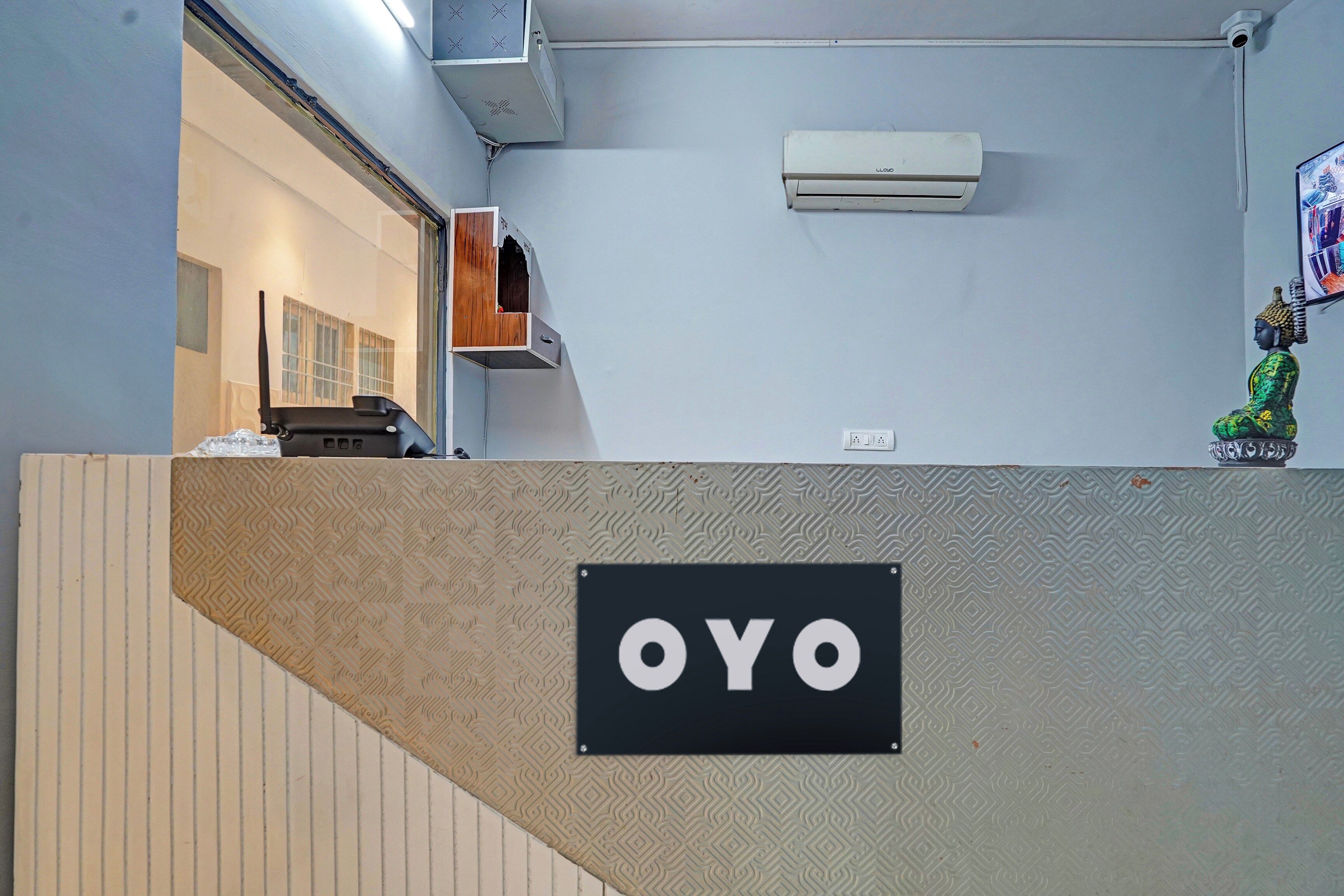 A reception area with a sign reading 'OYO' on a textured wall, an air conditioning unit mounted on the wall, and a small Buddha statue on the right.