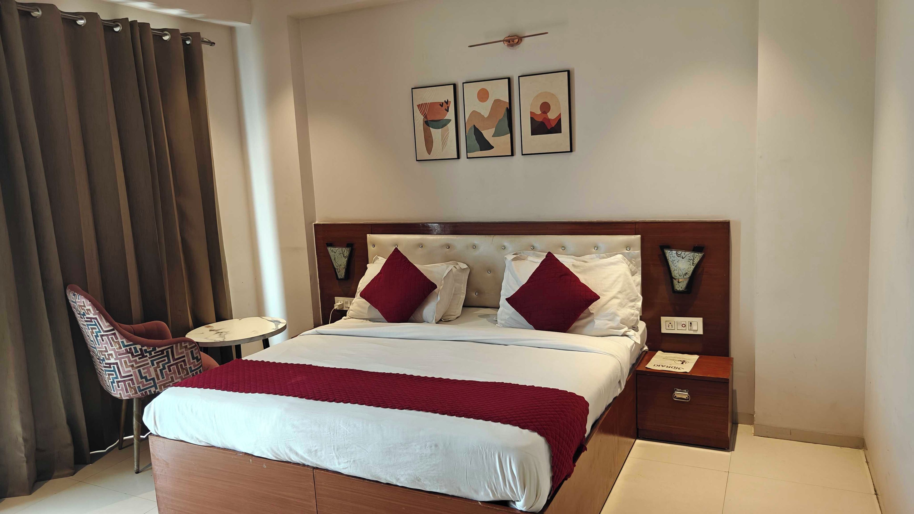 A hotel room with a bed, a chair, a small table, and artwork on the wall.