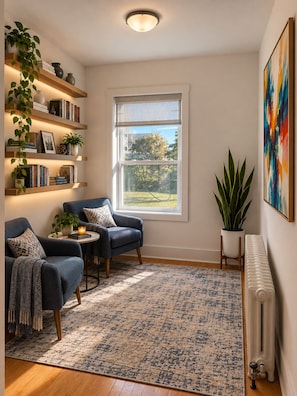 Una sala de estar acogedora con un conjunto de sofá azul, una mesa de centro y un tapete estampado. Hay estanterías con libros y objetos decorativos, y un ventanal con vistas al exterior.