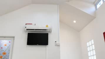 A room with a ceiling fan, an air conditioning unit, a flat-screen TV, a refrigerator, a wooden table, and a window.