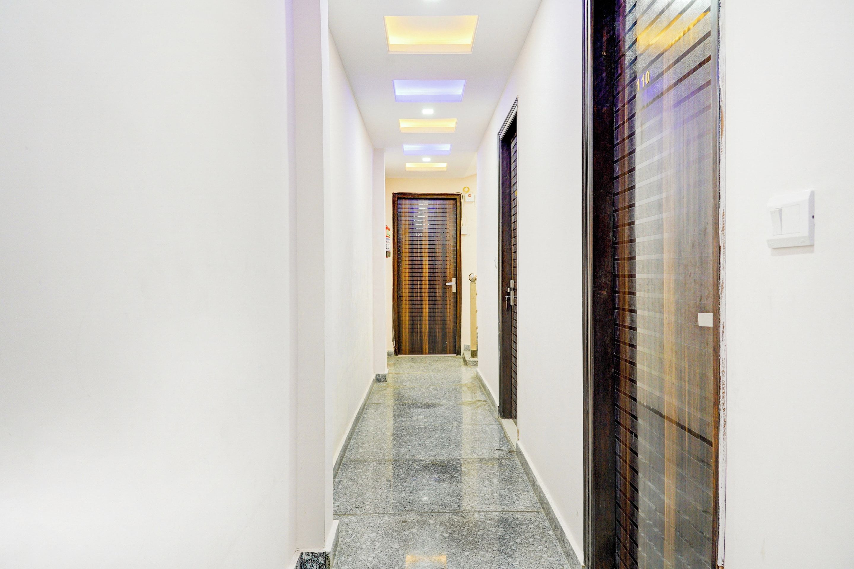 A hallway with a tiled floor, a wooden door at the end, and a wall-mounted light fixture.