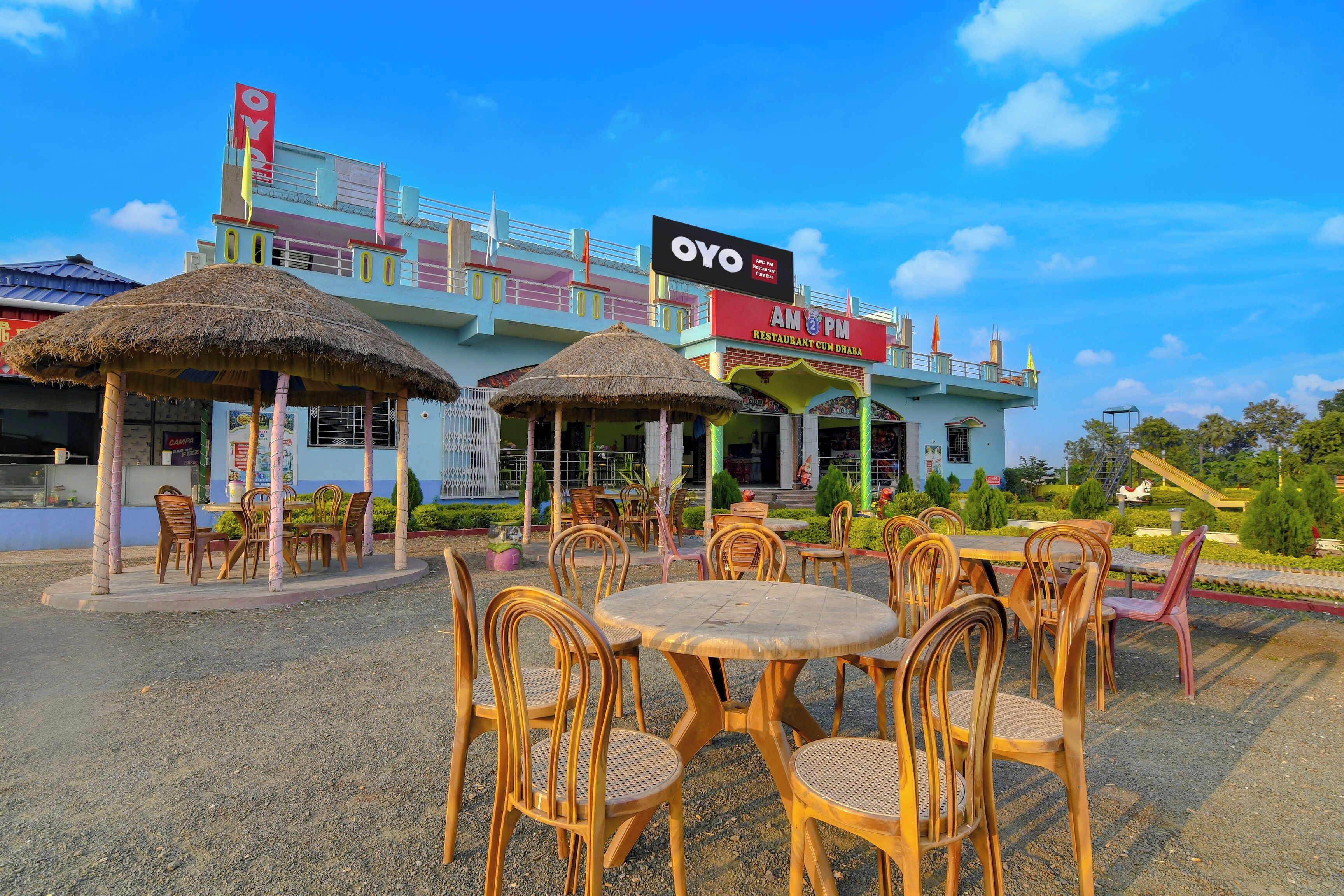 Un área de estar al aire libre con mesas y sillas redondas, una estructura con techo de paja y un edificio con fachada colorida y señalización para un restaurante y bar.