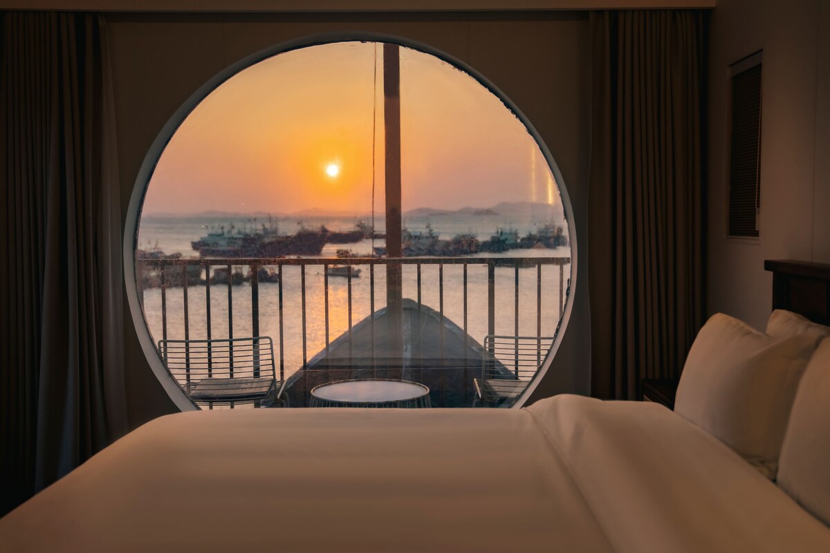A hotel room with a large circular window overlooking a body of water and a sunset.