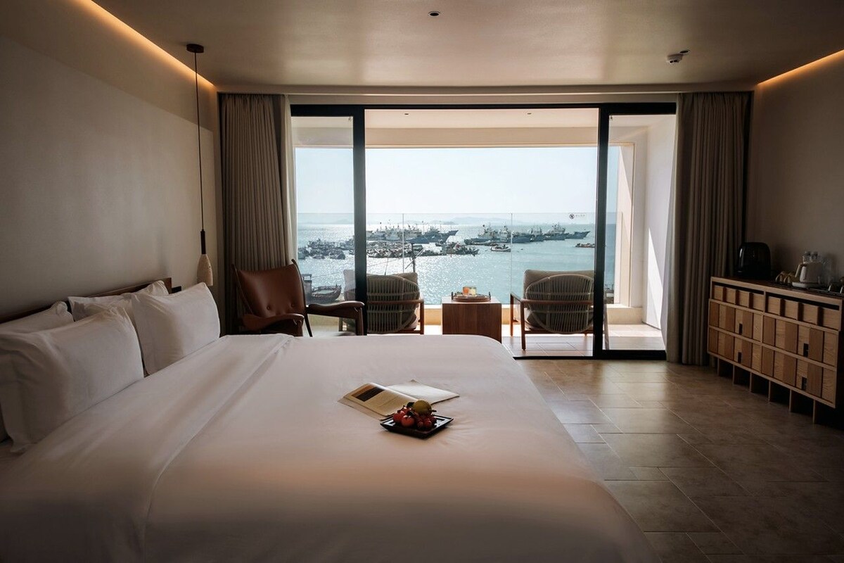 A hotel room with a large bed, a chair, a small table, and a view of the sea.