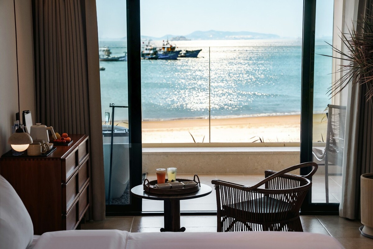 A hotel room with a large window offering a view of the beach and boats. There is a bed, a table with drinks, and a chair.