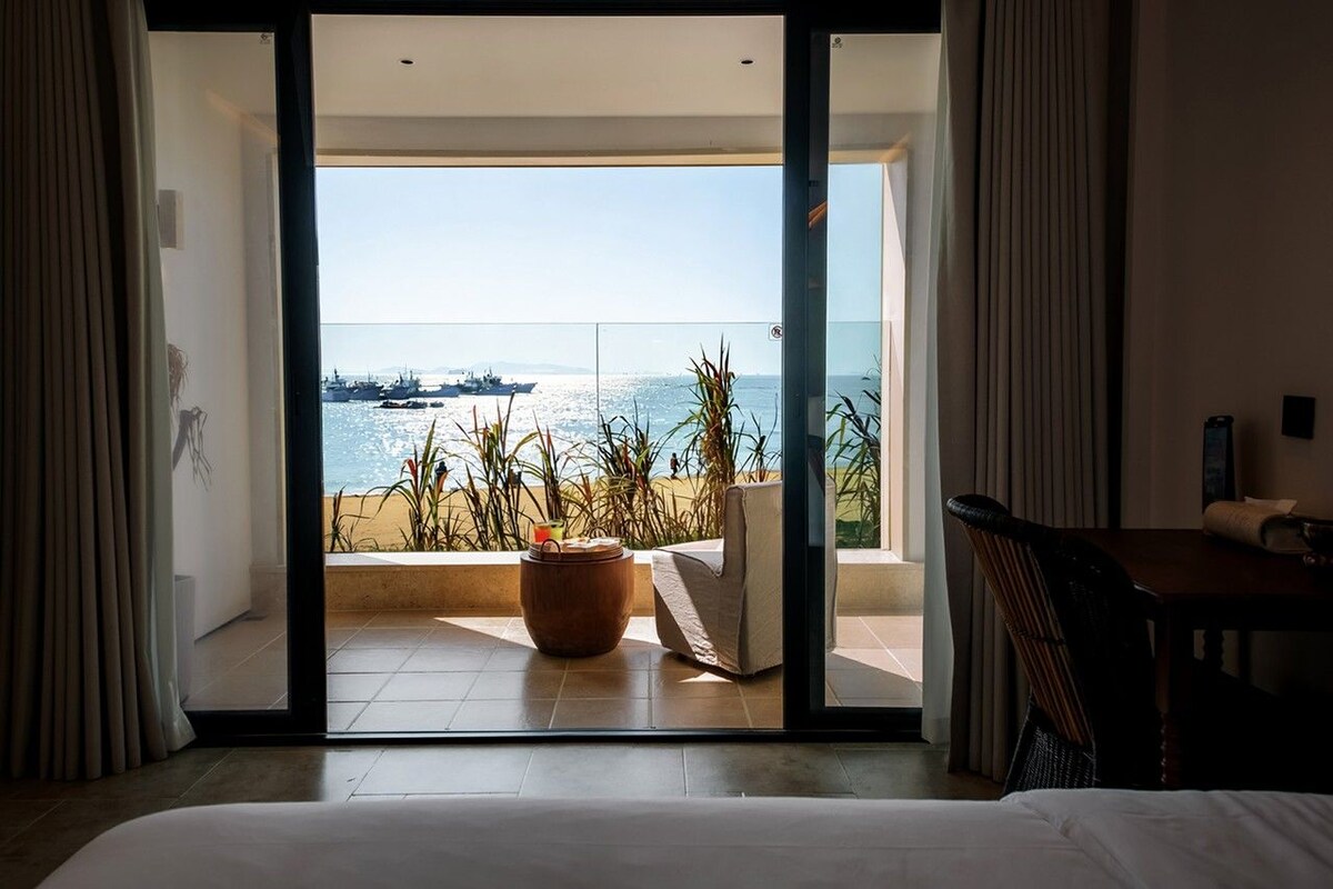 A hotel room with a large sliding glass door leading to a balcony with a view of the sea and boats.