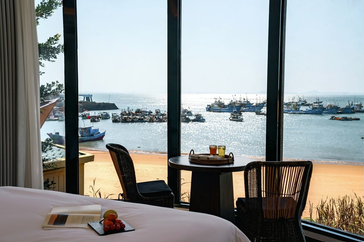 A hotel room with a large window overlooking a beach and a marina. There is a bed with a book and fruit on it, a wicker chair, and a small table with drinks.