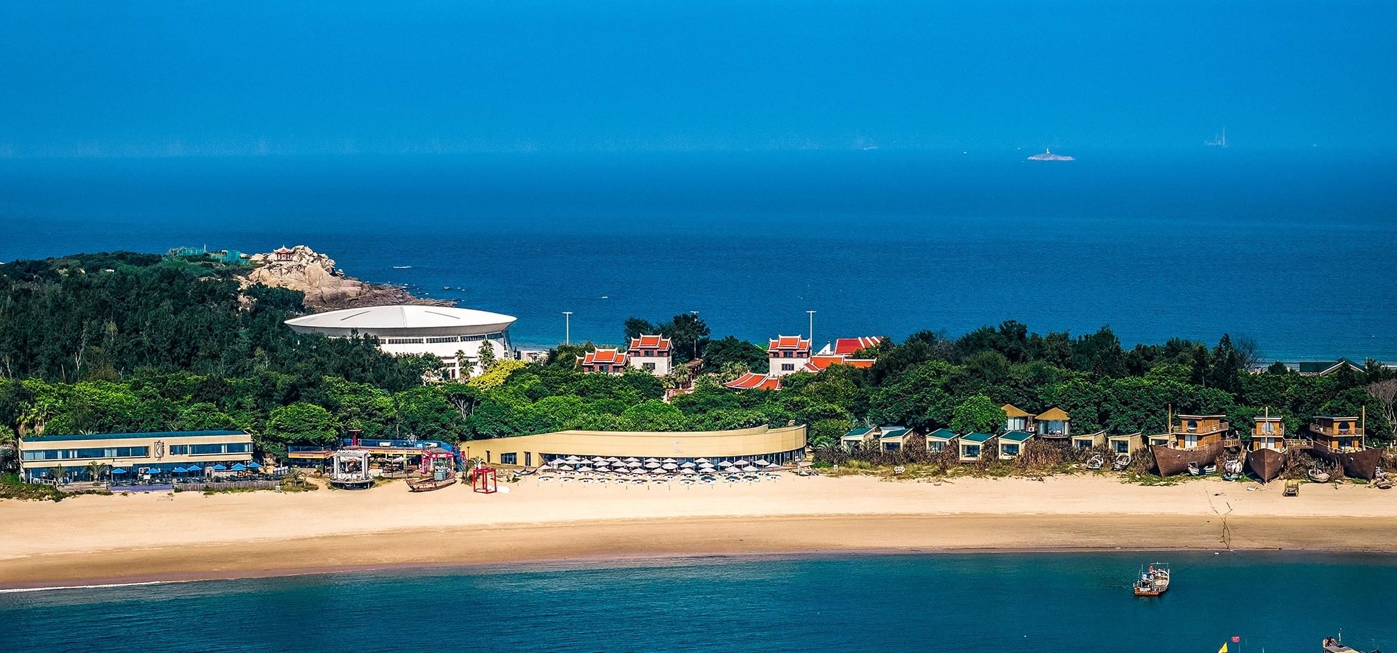 Un complejo turístico costero con playa, un edificio circular grande y varias construcciones más pequeñas.